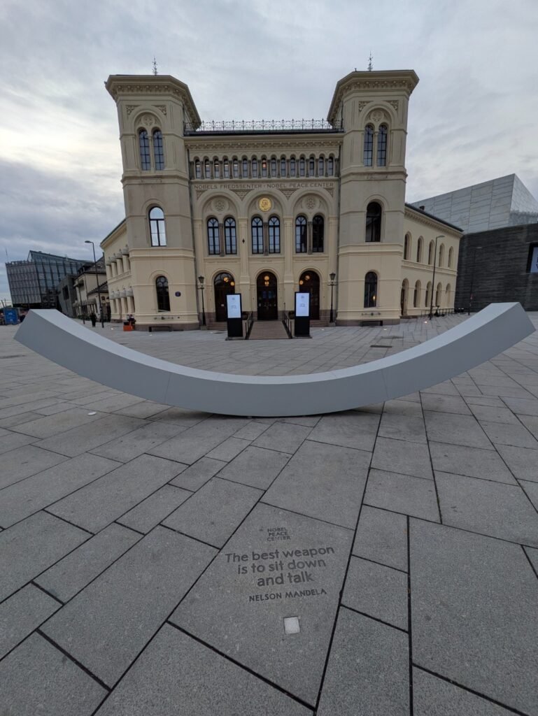 Il Nobel Peace Center di Oslo visto dalla piazza, con una scultura bianca a forma di sorriso e, incisa sulla pavimentazione, la frase di Nelson Mandela "The best weapon is to sit down and talk"